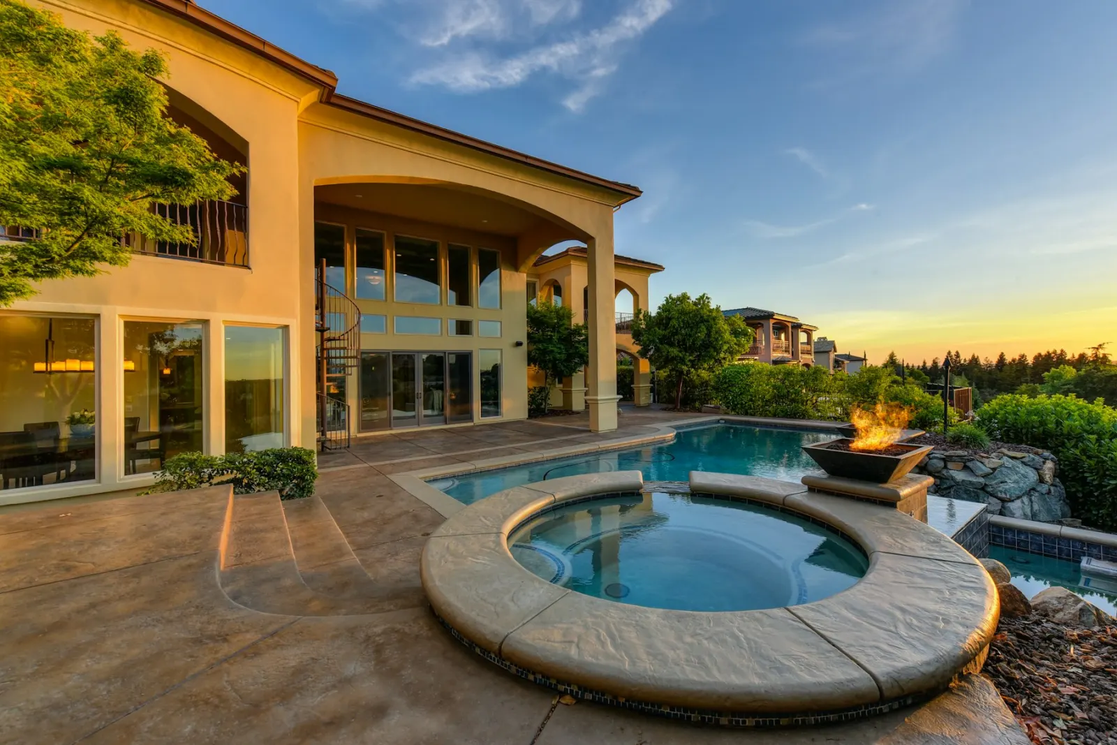 A house at sunset with a swimming pool, a round spa, and a lit fire bowl.