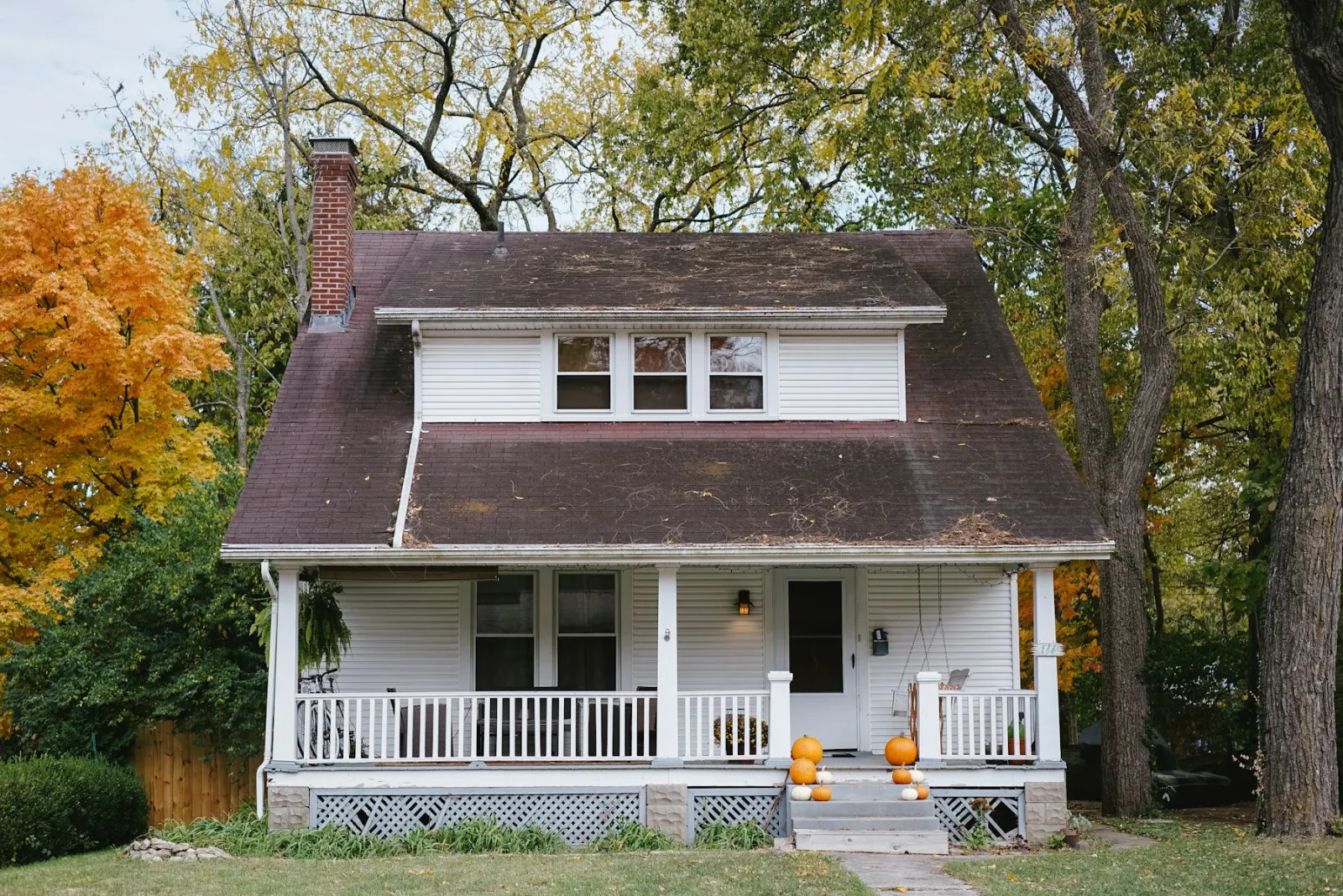 A white house with a covered porch, autumn trees, and pumpkins on the steps.